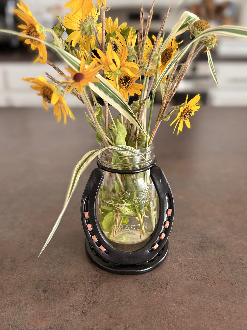 A small clear glass vase with a black deco base, filled with yellow daisy-like flowers and long green variegated leaves, placed on a brown countertop in a kitchen setting.